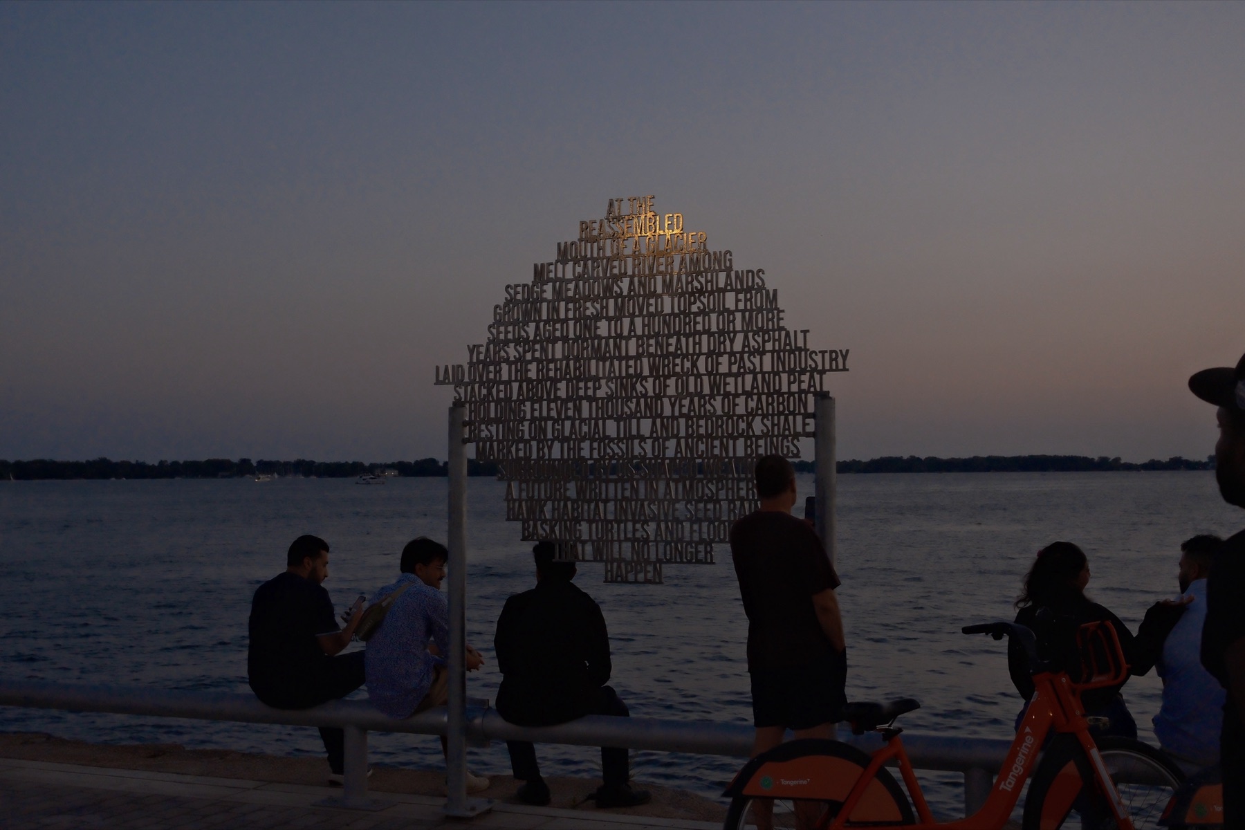 A poem cut from steel standing over the water at dusk, people on the railing