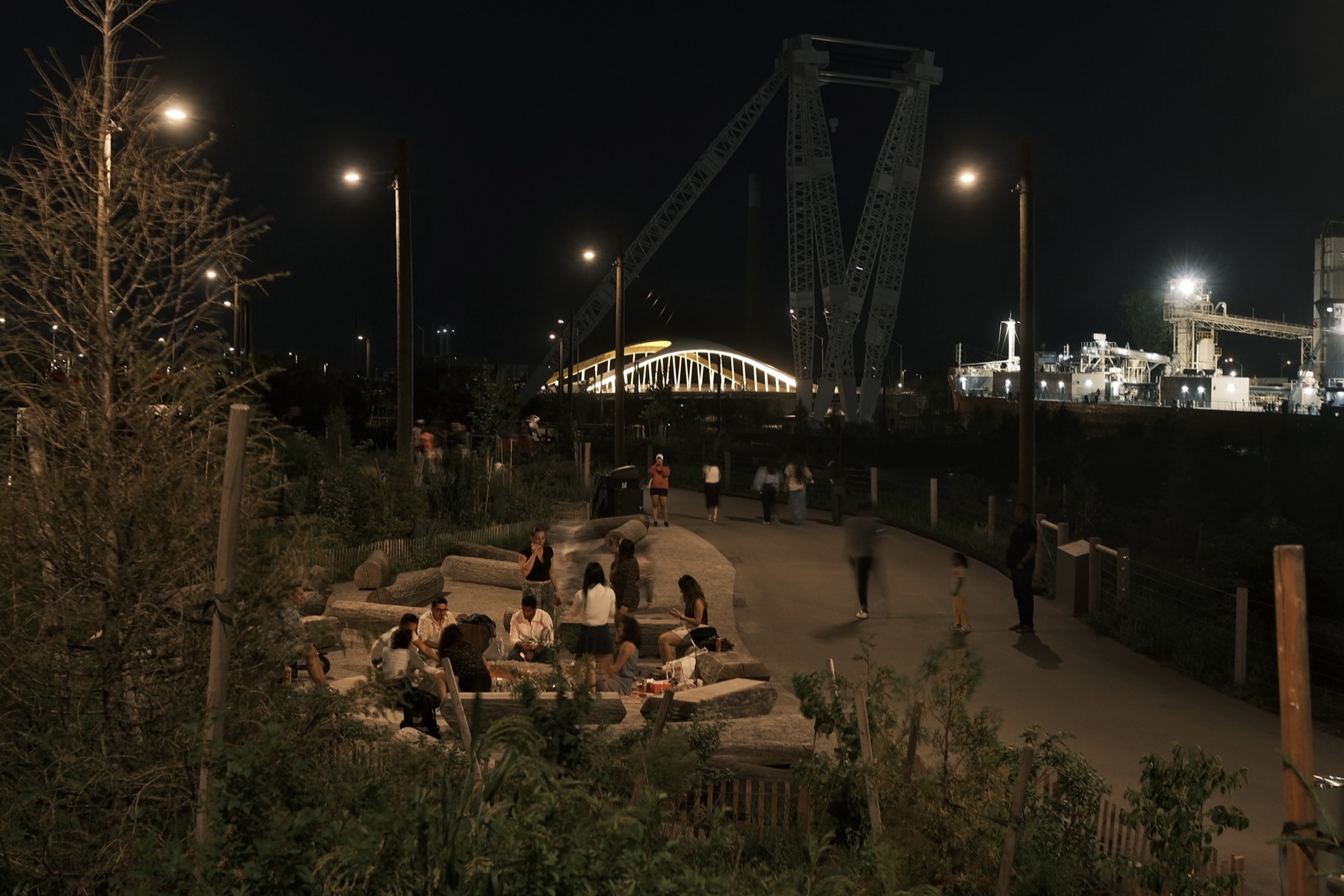 Groups at the log seating under the park lights, the white bridge lit, people walking off