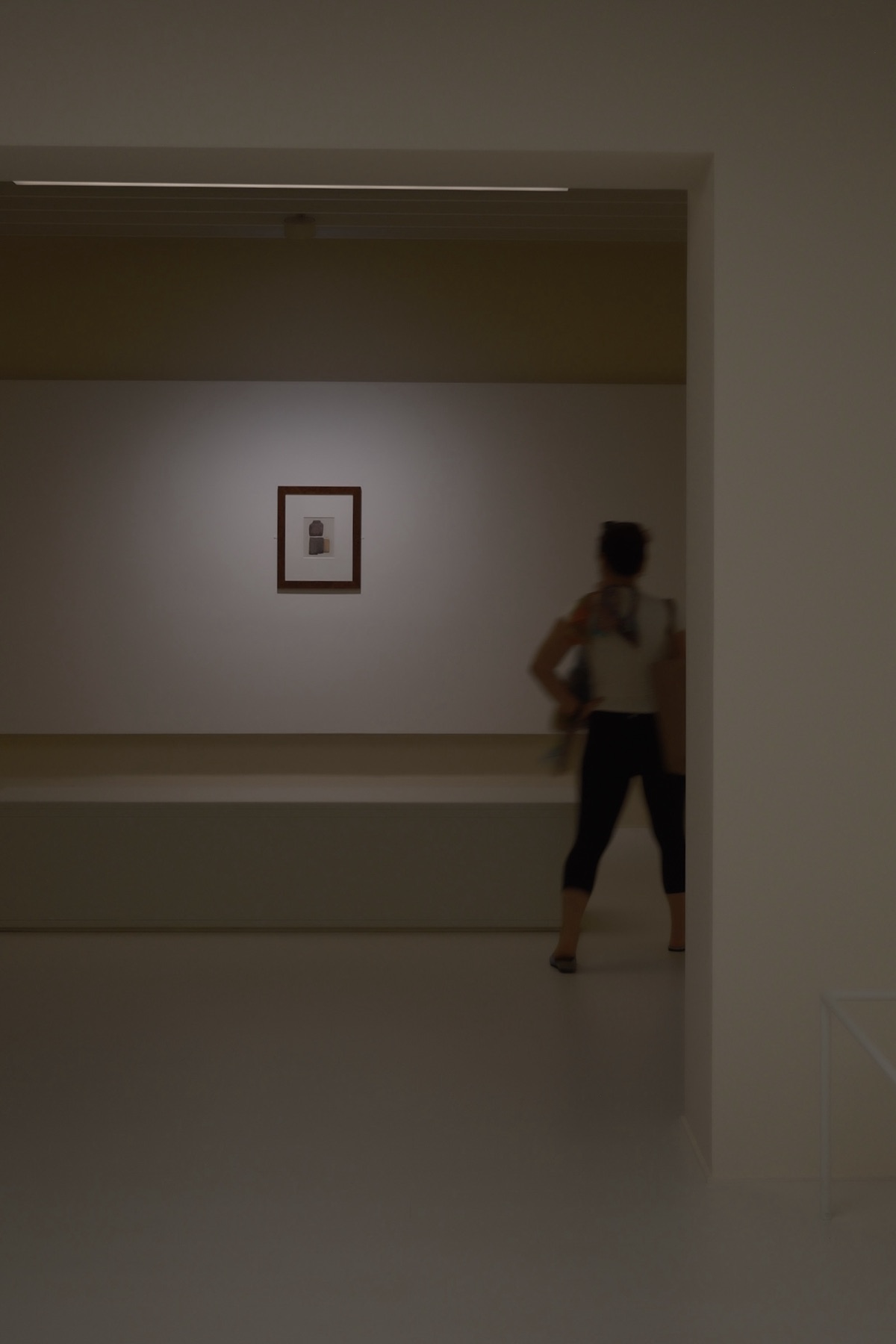 A single spotlit Morandi on a wide wall, a visitor walking out of frame — Still, After the Move