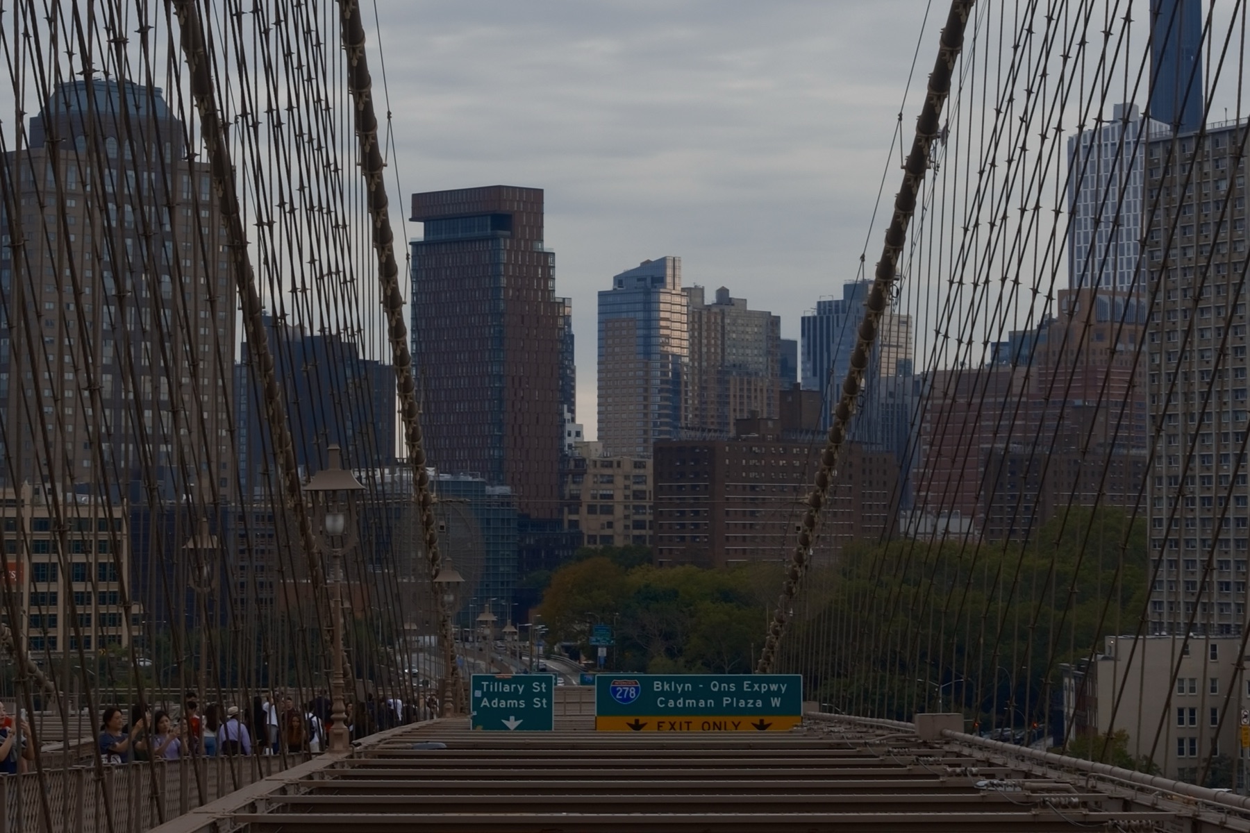 The bridge cables opening toward Brooklyn, green exit signs below