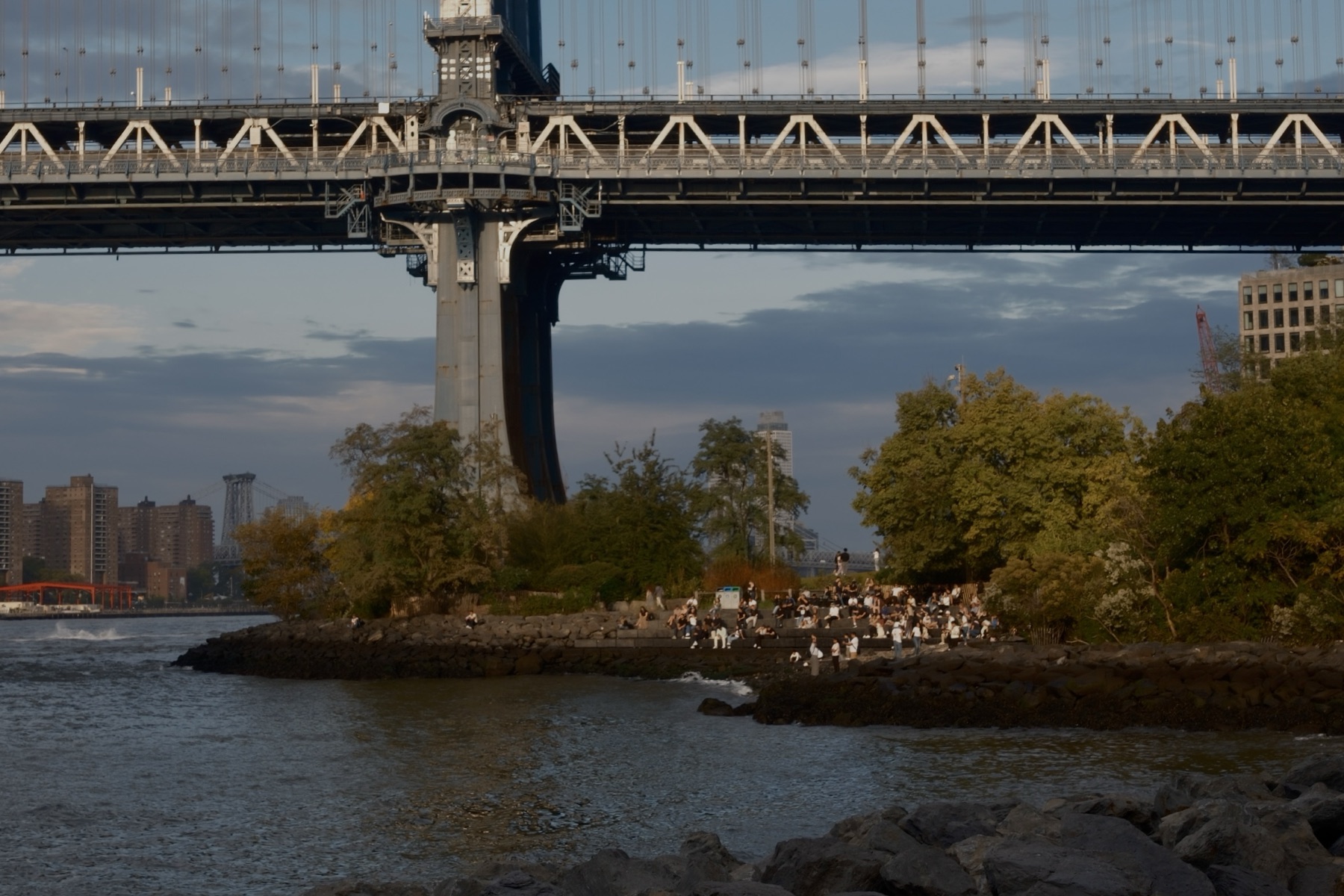 Rows of people on the stone steps under the Manhattan Bridge, lit by the low sun